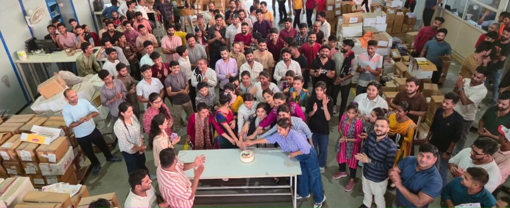 A large, diverse group of AAJ SCM employees gathered joyfully around a festive cake at a Reward and Recognition event, with many smiling and some gesturing towards the camera.